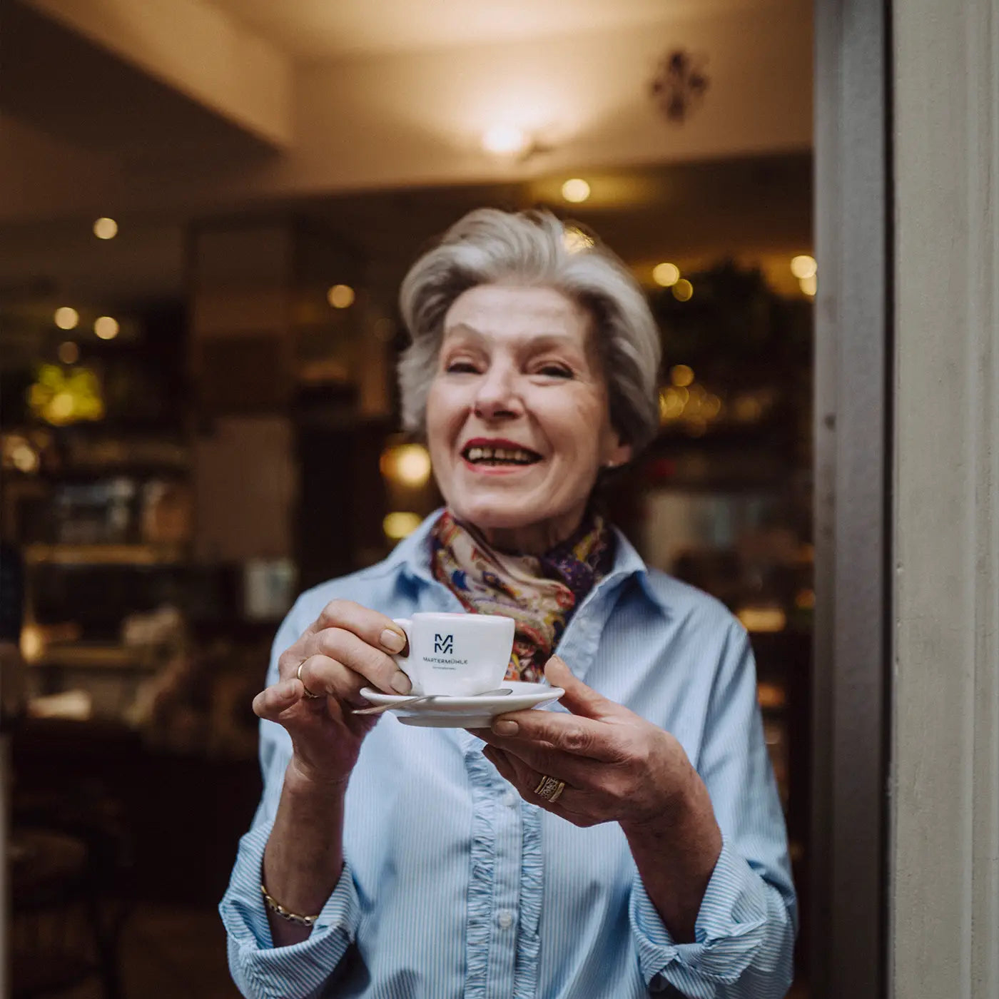 Eine alte lächelnde Frau am Eingang eines Cafés mit einer Espressotasse der Martermühle Kaffeerösterei