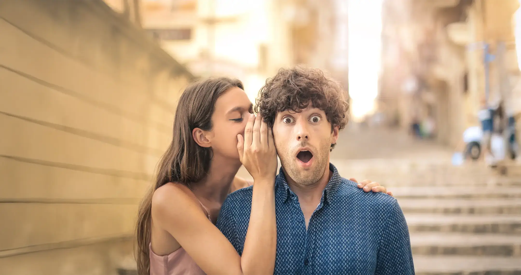 Junge Frau, die einem jungen Mann etwas ins Ohr flüstert.