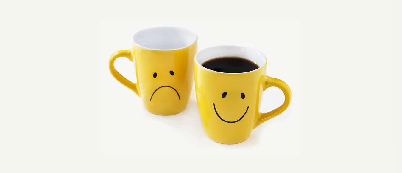 Zwei gelbe Tassen mit einem traurigen Smiley und einem lachenden Smiley, letztere ist gefüllt mit schwarzem Kaffee der Kaffeerösterei Martermühle