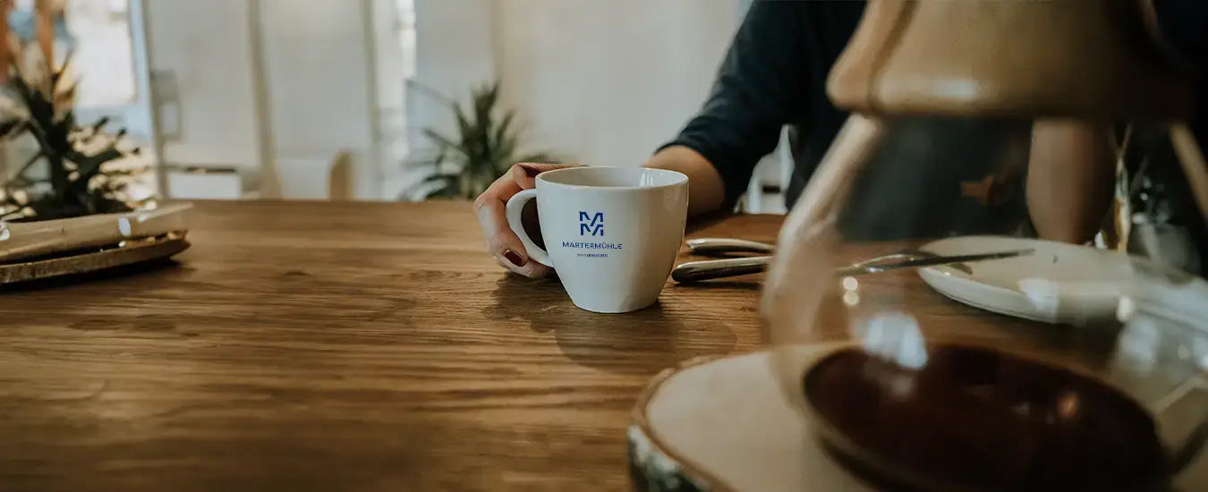 Eine Tasse der Kaffeerösterei Martermühle auf einem dunklen Holztisch und einer Chemex im Vordergrund
