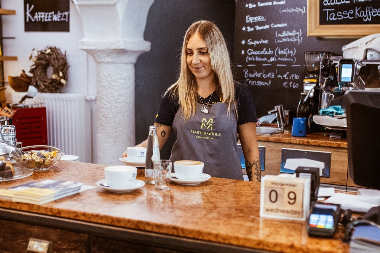 Eine Service Mitarbeiterin des Cafés der Kaffeerösterei in Assling hinter der Theke mit einer Tasse Cappuccino in der Hand